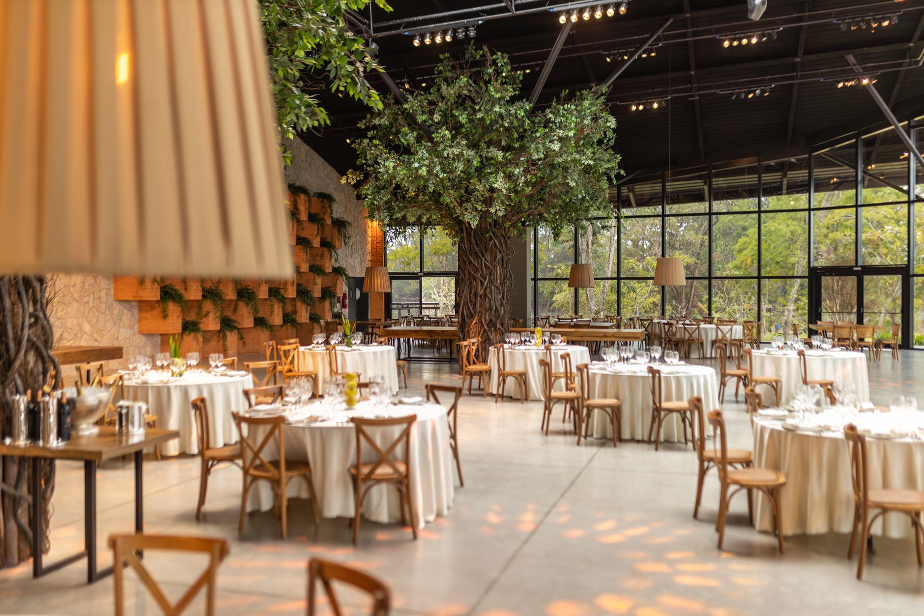 Spacious event hall featuring round tables, chairs, and indoor trees. Ideal for weddings or conferences. — Unlock the Hidden